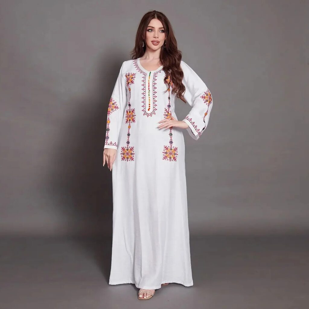 Femme debout en robe blanche kabyle à manches longues, ornée de broderies multicolores sur les manches et le torse